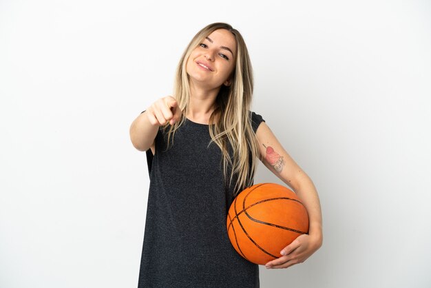 Junge Frau, die Basketball über isolierter weißer Wand spielt, die mit glücklichem Ausdruck nach vorne zeigt