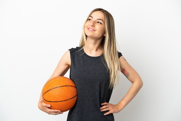 Junge Frau, die Basketball über isolierter weißer Wand spielt, posiert mit Armen an der Hüfte und lächelt