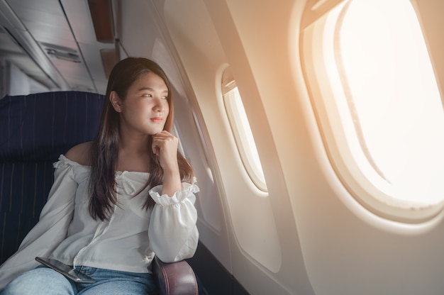 Junge Frau, die bei Sonnenuntergang in der Nähe des Flugzeugfensters sitzt und während des Fluges Handy benutzt