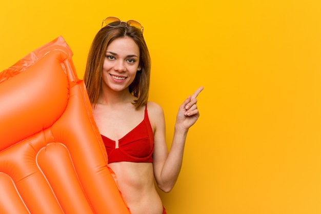 Junge Frau, die Bikini trägt und ein Luftmatratzebett hält, das fröhlich mit Zeigefinger weg zeigt.