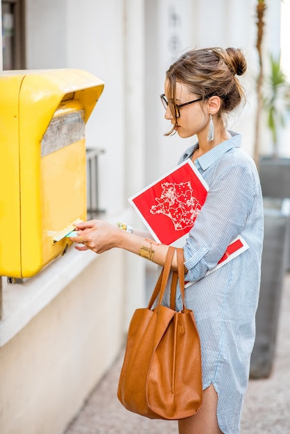 Junge Frau, die Brief an den alten gelben Briefkasten legt, der mit Tasche und Zeitschrift draußen auf der Straße steht