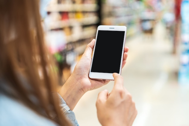 Junge Frau, die das Smartphone des leeren Bildschirms beim Einkaufen im Lebensmittelgeschäft am Supermarkt hält