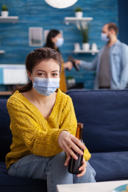 Junge Frau, die die Kamera betrachtet, die im Wohnzimmer zu Hause soziale Distanz hält und im Zuge der globalen Pandemie mit Coronavirus eine Gesichtsmaske trägt, die eine Bierflasche hält.