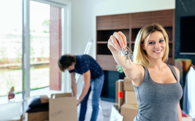 Junge Frau, die die Schlüssel ihres neuen Hauses zeigt, während ihr Mann Kisten auspackt Selektiver Fokus auf Schlüssel im Vordergrund