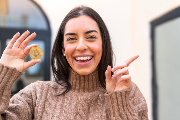 Junge Frau, die draußen einen Bitcoin hält, überrascht und zeigt mit dem Finger zur Seite