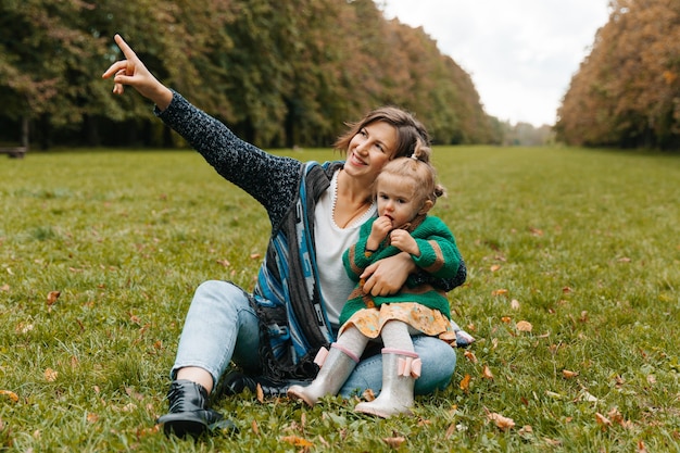 Junge Frau, die ein Baby in ihren Armen hält und auf dem Gras im Park sitzt. Mama und Tochter gehen im Park spazieren.