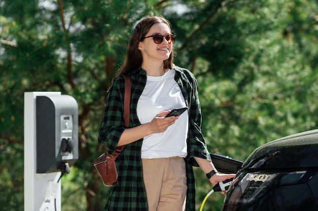 Junge Frau, die ein Elektroauto auflädt und beim Aufladen auf dem Smartphone im Internet surft