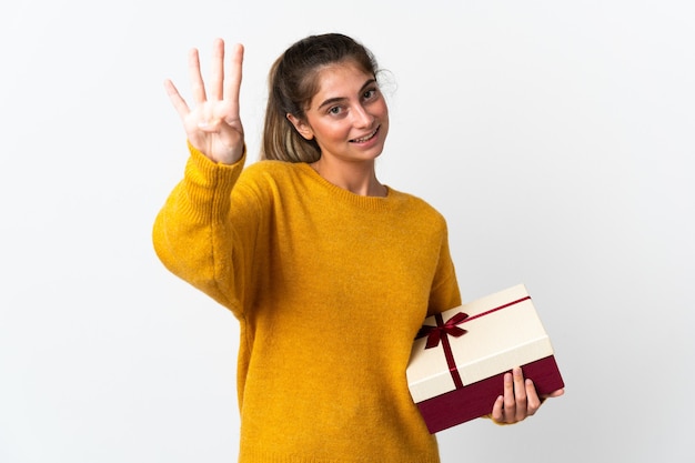 Junge Frau, die ein Geschenk lokalisiert auf weißer Wand glücklich hält und vier mit den Fingern zählt