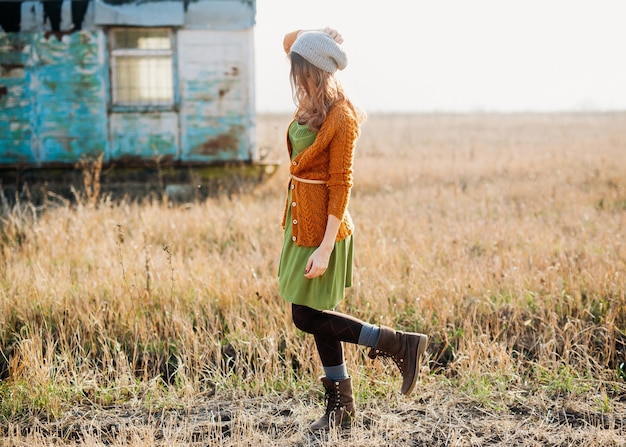 Junge Frau, die ein Kleid, eine gestrickte Strickjacke und Stiefel trägt und auf einem Feld geht