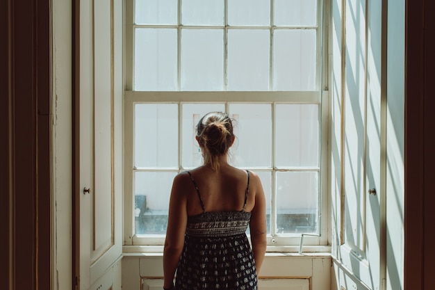 Junge Frau, die ein Kleid trägt, das durch ein Fenster schaut, der Kamera den Rücken gibt, Stress- und Angstkonzepte, dunkles Bild, Traurigkeit