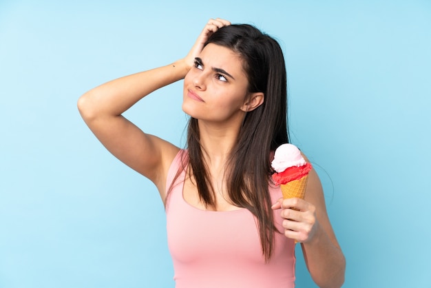 Junge Frau, die ein Kornett-Eis über isoliertem Blau hält, das Zweifel und mit verwirrendem Gesichtsausdruck hat