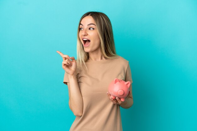 Junge Frau, die ein Sparschwein über einer isolierten blauen Wand hält, die beabsichtigt, die Lösung zu realisieren, während sie einen Finger hochhebt