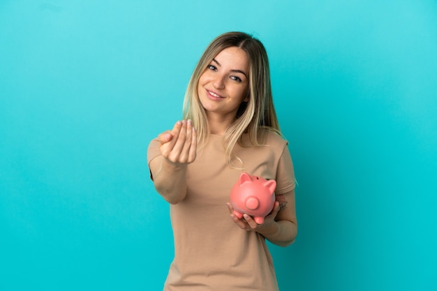 Junge Frau, die ein Sparschwein über isoliertem blauem Hintergrund hält und einlädt, mit der Hand zu kommen. Schön, dass du gekommen bist