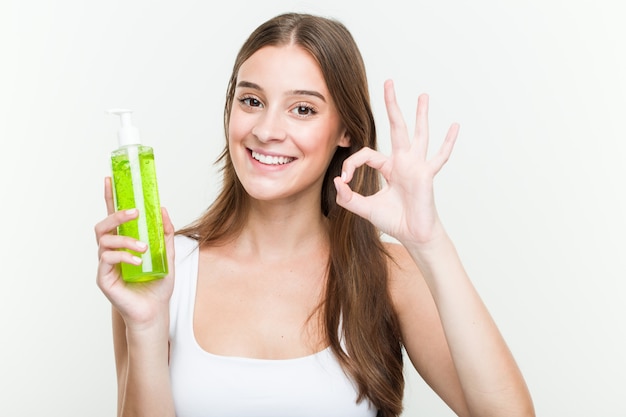 Junge Frau, die eine Aloe Vera-Flasche nett und überzeugt hält, okaygeste zeigend