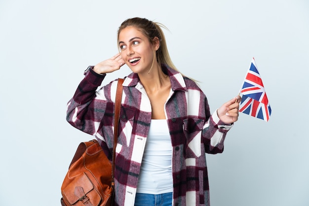 Junge Frau, die eine britische Flagge lokalisiert auf weißem Hintergrund hält, der etwas hört, indem man Hand auf das Ohr setzt