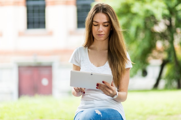 Junge Frau, die eine digitale Tablette sitzt auf einer Banch in einem Park verwendet