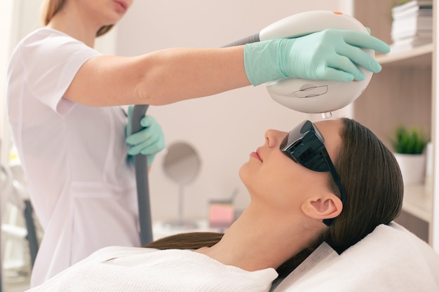 Junge Frau, die eine dunkle Brille trägt, während sie auf der medizinischen Couch liegt, und eine Kosmetikerin, die ein Lasergerät über ihrer Stirn hält