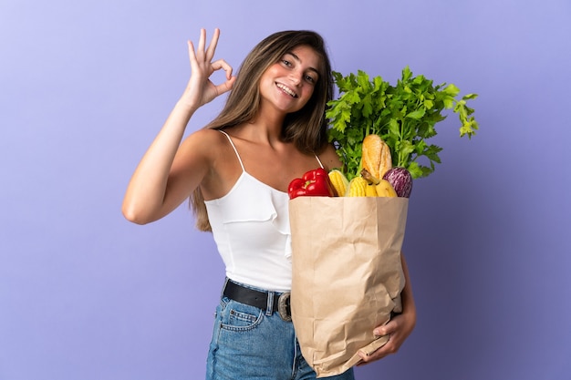 Junge Frau, die eine Einkaufstasche des Lebensmittels hält ok Zeichen mit den Fingern zeigt