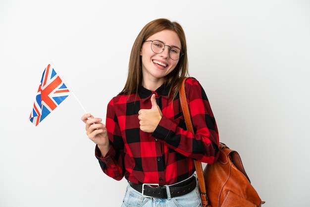 Junge Frau, die eine Flagge des Vereinigten Königreichs isoliert auf blauem Hintergrund hält und eine Geste mit dem Daumen nach oben gibt