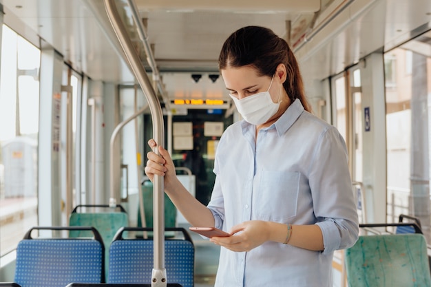 Junge Frau, die eine Gesichtsmaske trägt und am Telefon während des Reisens mit öffentlichen Verkehrsmitteln plaudert