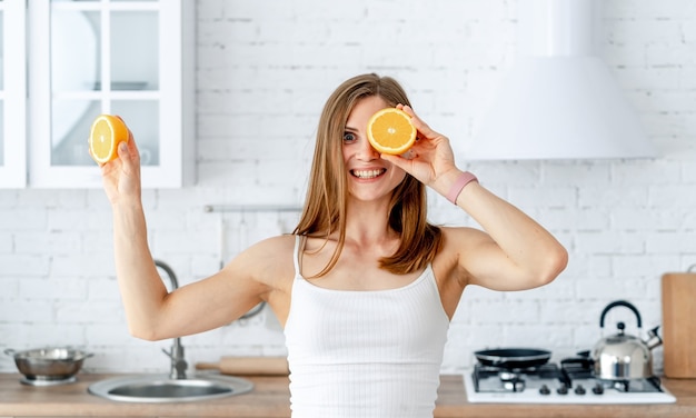 Junge Frau, die eine Hälfte der Orange nahe ihrem schönen Gesicht im Küchenhintergrund hält. Gesundes Lebensmittelkonzept.