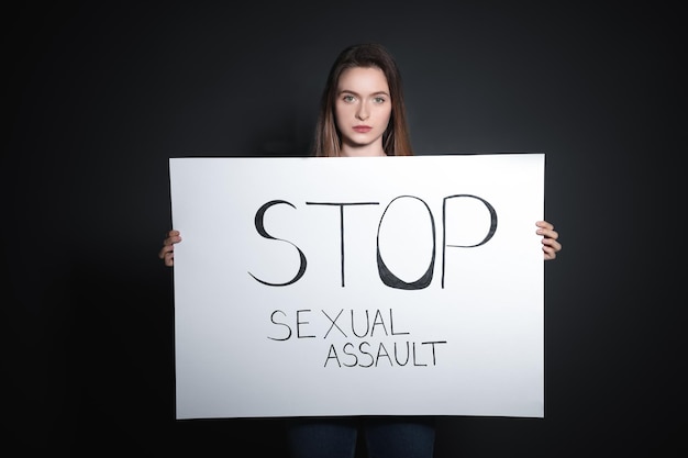 Junge Frau, die eine Karte mit den Worten STOPP SEXUELLEN ÜBERGRIFF vor dunklem Hintergrund hält