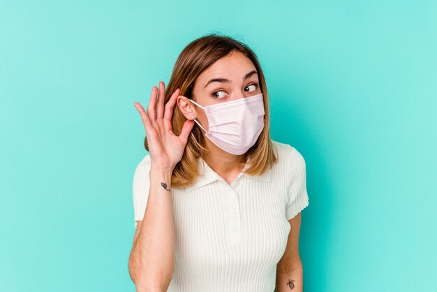 Junge Frau, die eine Maske für Virus lokalisiert auf blauer Wand trägt, die versucht, einen Klatsch zu hören.