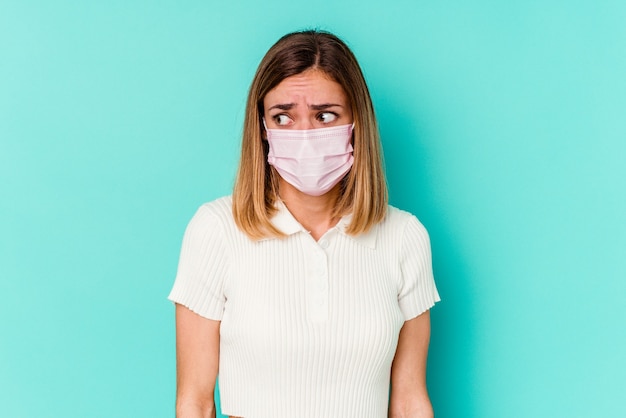 Junge Frau, die eine Maske für Virus trägt, die auf blauer Wand verwirrt ist, fühlt sich zweifelhaft und unsicher