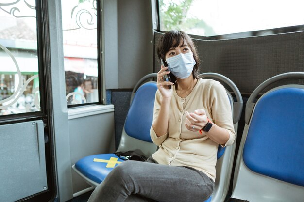 Junge Frau, die eine Maske trägt, sitzt auf einer Bank, während sie unterwegs im Bus telefoniert