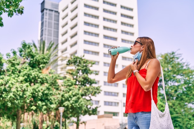 Junge Frau, die eine Maske Trinkwasser in der Stadt trägt