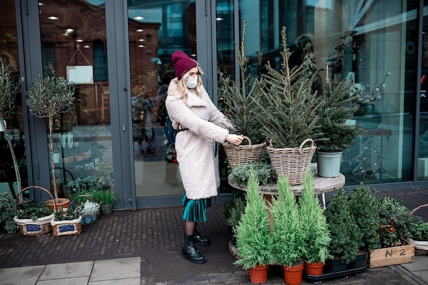 Junge Frau, die eine medizinische Schutzmaske in Kunstpelzmantel und Hut trägt, pflückt kleinen Weihnachtsbaum im Korbtopf im Blumenladen der Stadt. Weihnachtsmarkt, Vorbereitung auf den Urlaub