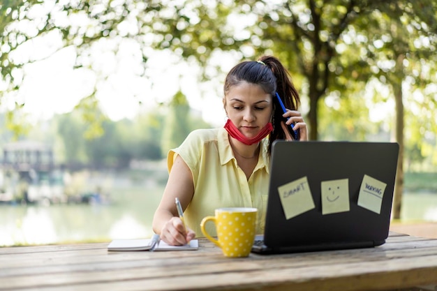 Junge Frau, die eine offene Schutzmaske mit Laptop im Freien trägt Unternehmerin, die sich Notizen macht, während sie mit dem Handy im Park anruft Fernarbeit neues normales Konzept nach der Covid-19-Pandemie