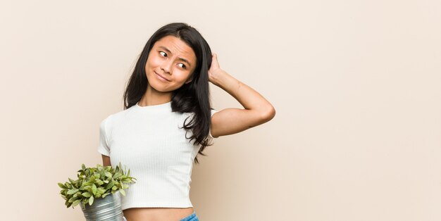 Junge Frau, die eine Pflanze hält, die Hinterkopf berührt, denkt und eine Wahl trifft.