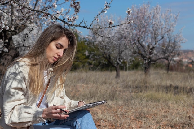Junge Frau, die eine Skizze in einem Notizbuch auf einem Baumgebiet im Freien zeichnet