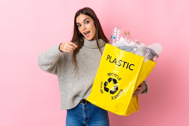 Junge Frau, die eine Tasche voll von Plastikflaschen hält, um isoliert auf rosa Wand überrascht und nach vorne zeigend zu recyceln