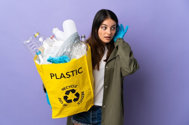 Junge Frau, die eine Tasche voller Plastikflaschen hält, um lokalisiert auf lila Wand zu recyceln, die etwas hört, indem man Hand auf das Ohr legt