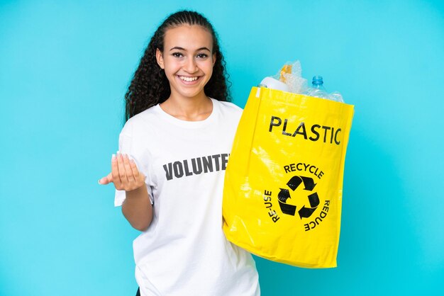 Junge Frau, die eine Tüte voller Plastikflaschen hält, um isoliert auf blauem Hintergrund zu recyceln, die einlädt, mit der Hand zu kommen Glücklich, dass Sie gekommen sind