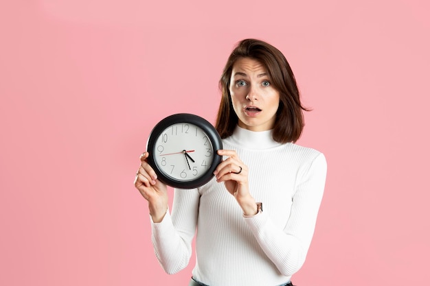 Junge Frau, die eine Uhr auf einem rosa Hintergrund hält