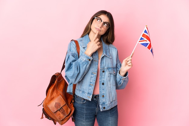 Junge Frau, die eine Vereinigte Königreichsflagge lokalisiert auf Rosa hält, die Zweifel beim Aufblicken hat