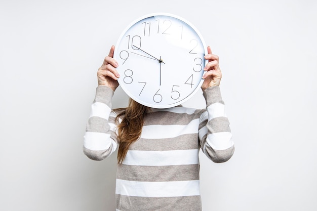 Junge Frau, die eine Wanduhr hält, die ihr Gesicht auf hellem Hintergrund bedeckt