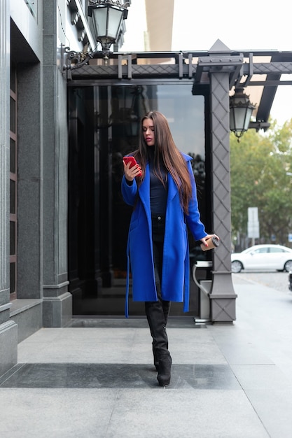 Junge Frau, die einen blauen Mantel trägt, trinkt Kaffee zum Mitnehmen und telefoniert auf der Straße. Gesperrter Echtzeit-Mittelschuss