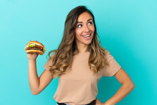 Junge Frau, die einen Burger über isoliertem Hintergrund hält und eine Idee denkt, während sie nach oben schaut