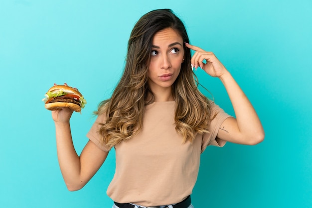 Junge Frau, die einen Burger über isoliertem Hintergrund hält und Zweifel hat und nachdenkt