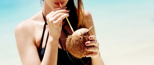 Junge Frau, die einen Cocktail an einem schönen Strand mit türkisfarbenem Wasser genießt.