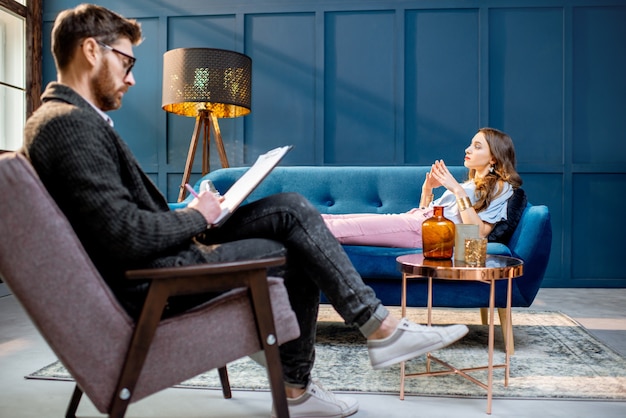 Junge Frau, die einen männlichen Psychologen besucht, der während einer psychologischen Sitzung im luxuriösen blauen Bürointerieur auf der bequemen Couch liegt