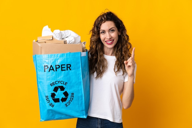 Junge Frau, die einen Recyclingbeutel voll Papier hält, um isoliert auf gelber Wand zu recyceln, die einen Finger im Zeichen des Besten zeigt und hebt