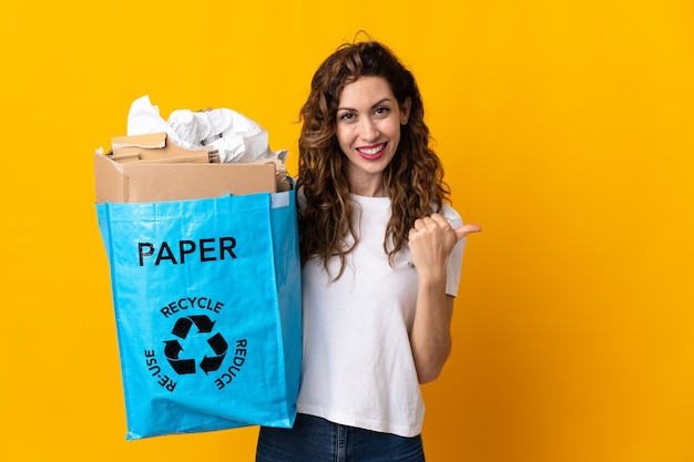 Junge Frau, die einen Recyclingbeutel voll Papier hält, um isoliert auf gelber Wand zu recyceln, die zur Seite zeigt, um ein Produkt zu präsentieren