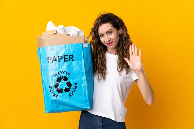 Junge Frau, die einen Recyclingbeutel voll Papier hält, um lokalisiert auf gelber Wand, die mit Hand mit glücklichem Ausdruck salutiert, zu recyceln