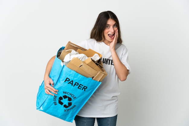 Junge Frau, die einen Recyclingbeutel voll Papier hält, um lokalisiert auf weißer Wand mit Überraschung und schockiertem Gesichtsausdruck zu recyceln