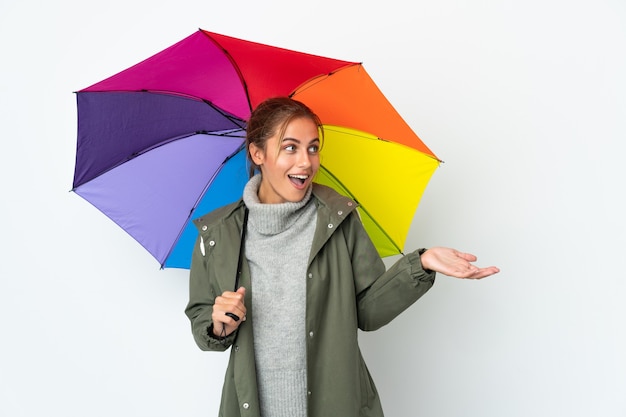 Junge Frau, die einen Regenschirm auf weißem Hintergrund mit Überraschungsausdruck beim Betrachten der Seite hält